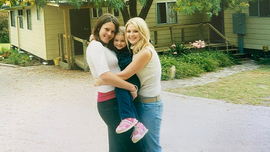 Two women holding a girl in front of a cabin