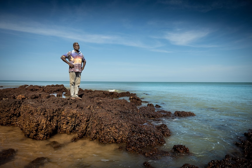 A man with his hands on his hips standing on a rocky outcrop on a beach, the ocean behind him.