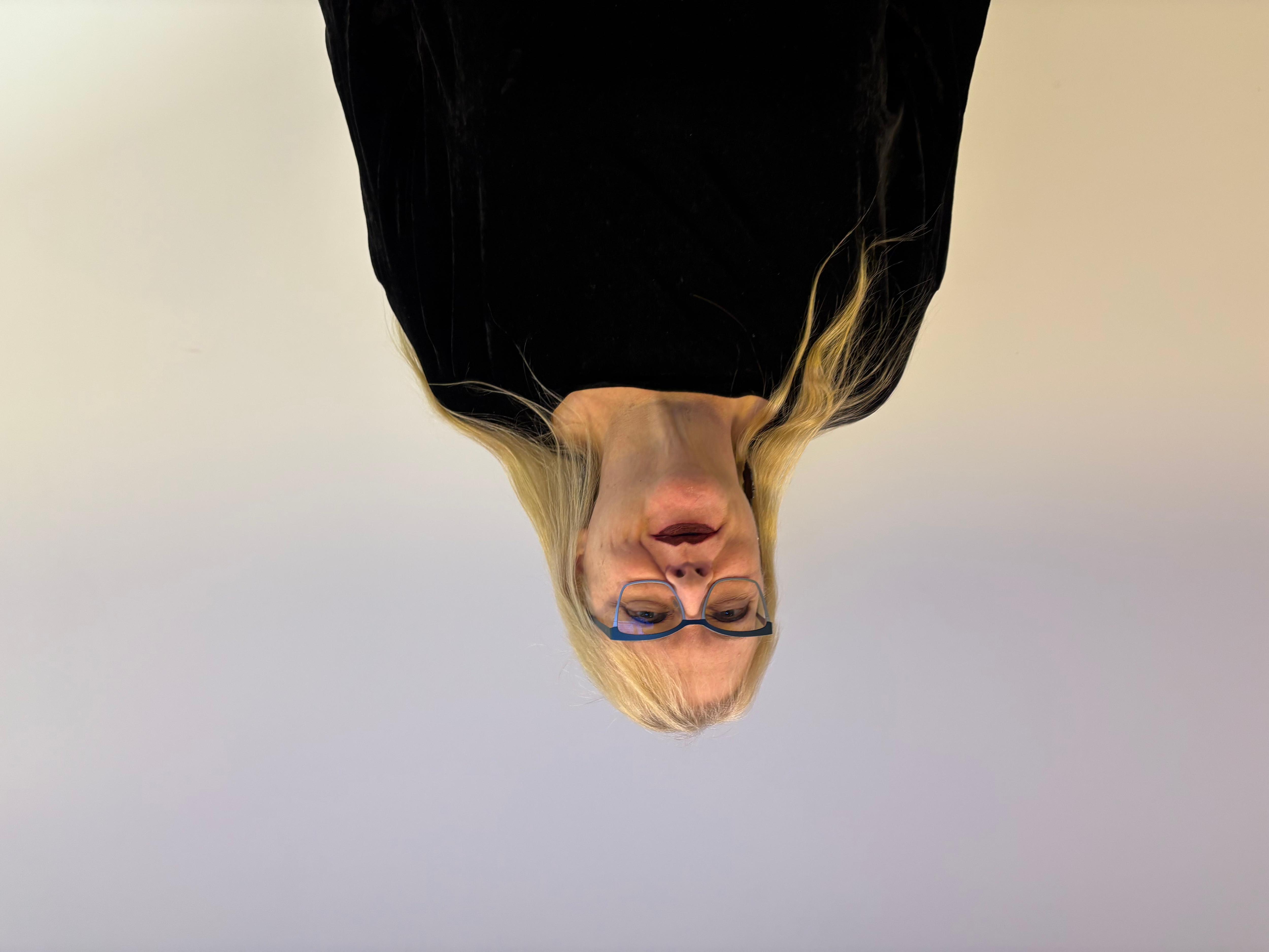 A woman with long blonde hair, blue framed glasses and a black top stands in a studeio with pastel purple and cream colours.