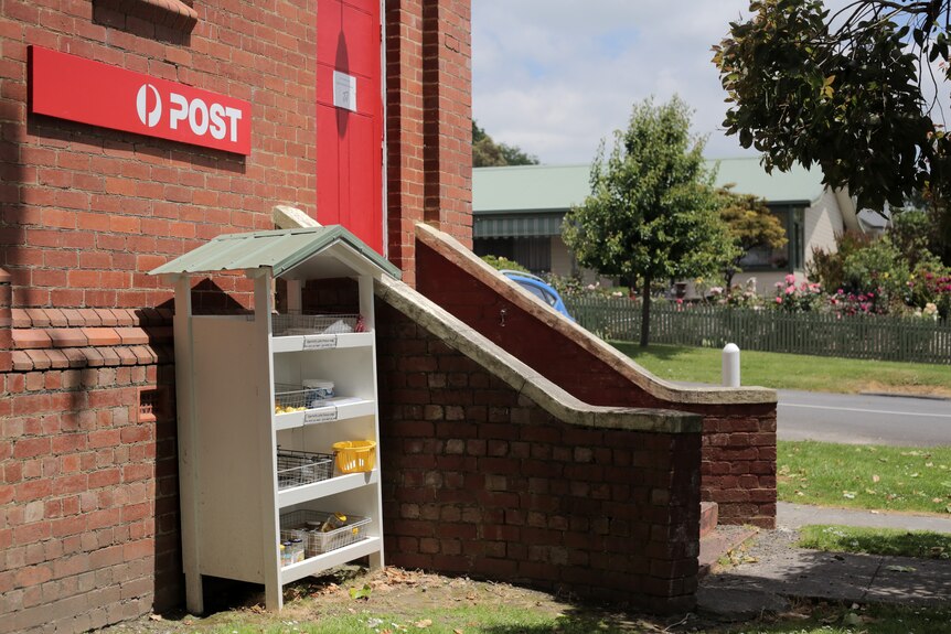 Exterior of a brick building with steps leading to a red door. 'Post' sign is above a white bookcase with roof.