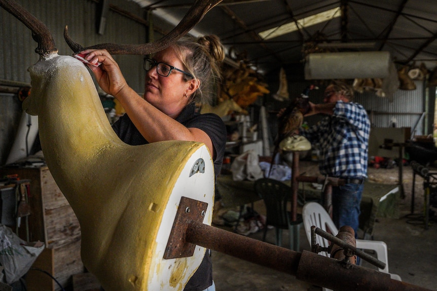 A woman in a black polo shirt sands a deer head mold in a large shed.