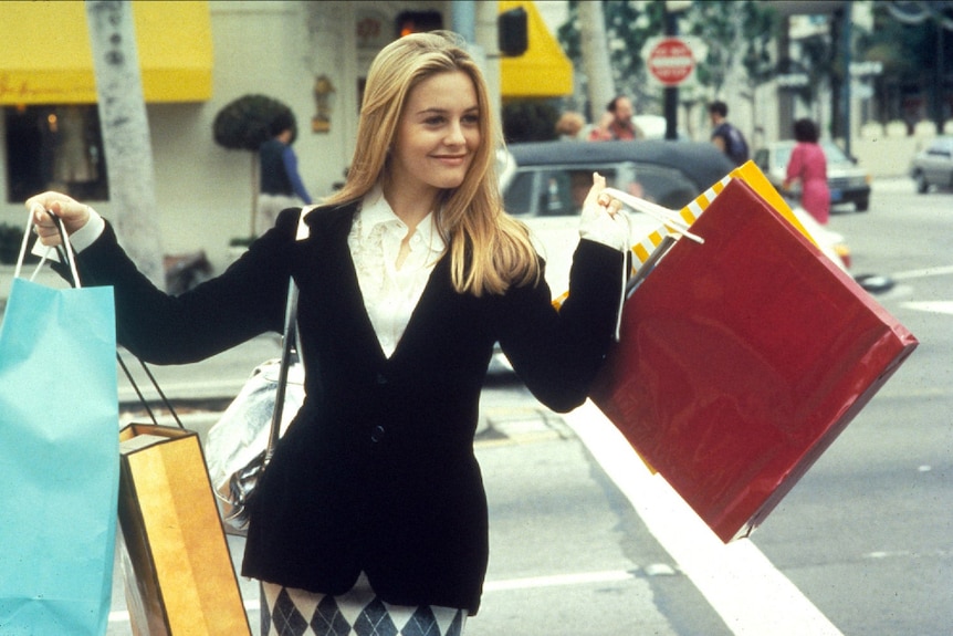 A happy young blonde woman in patterned skirt, white shirt and black blazer holds up handful of shopping bags on city street.