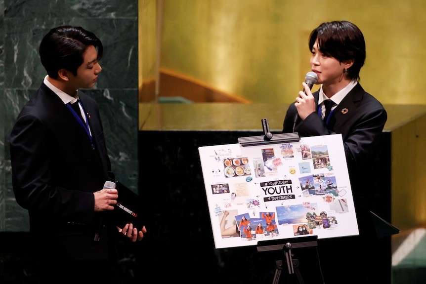 BTS member Jimin speaks into a microphone at the United Nations as bandmate Jungkook listens. They stand next to a collage.