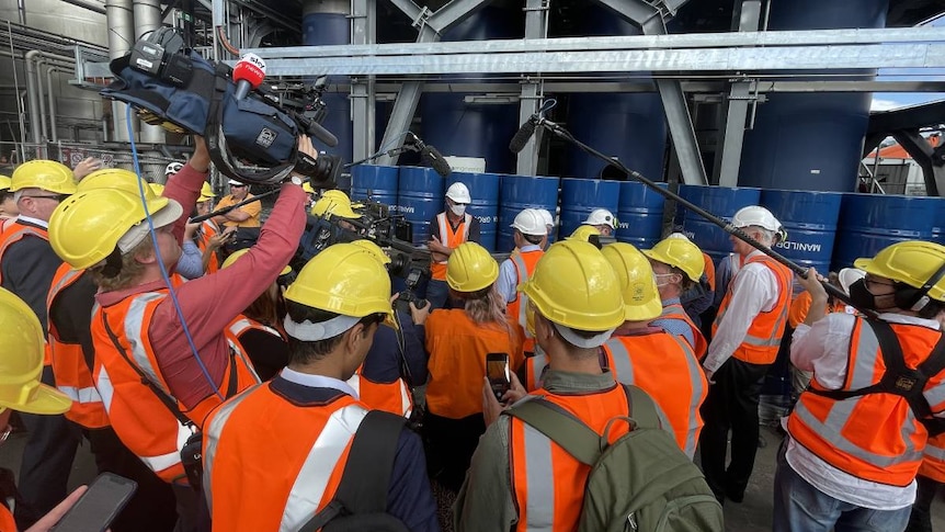 people gathering around in high viz with cameras and microphones at a manufacturing plant.