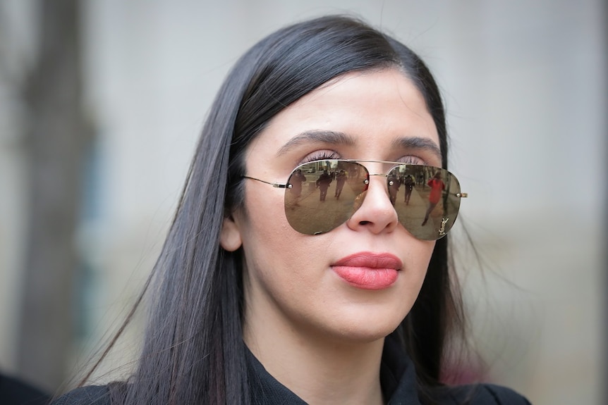 A woman wearing sunglasses looks straight ahead as she is photographed by media, who are reflected in her glasses.
