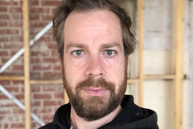 Chris pose pour un selfie sur un chantier, vêtu d'un sweat à capuche noir.