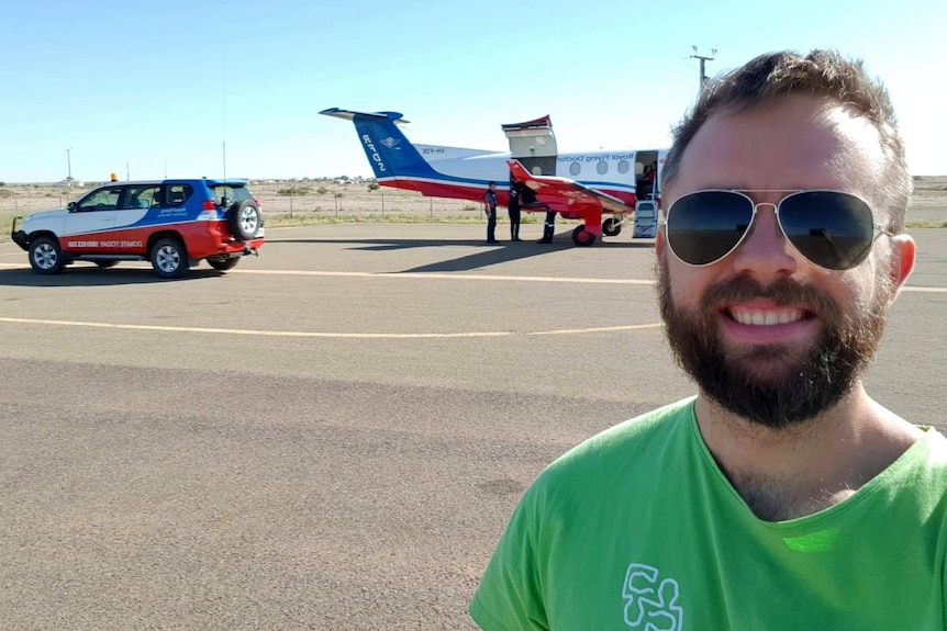 A man wearing a green shirt and black sunglasses with brown hair and a beard smiles at the camera in front of an RFDS plane.