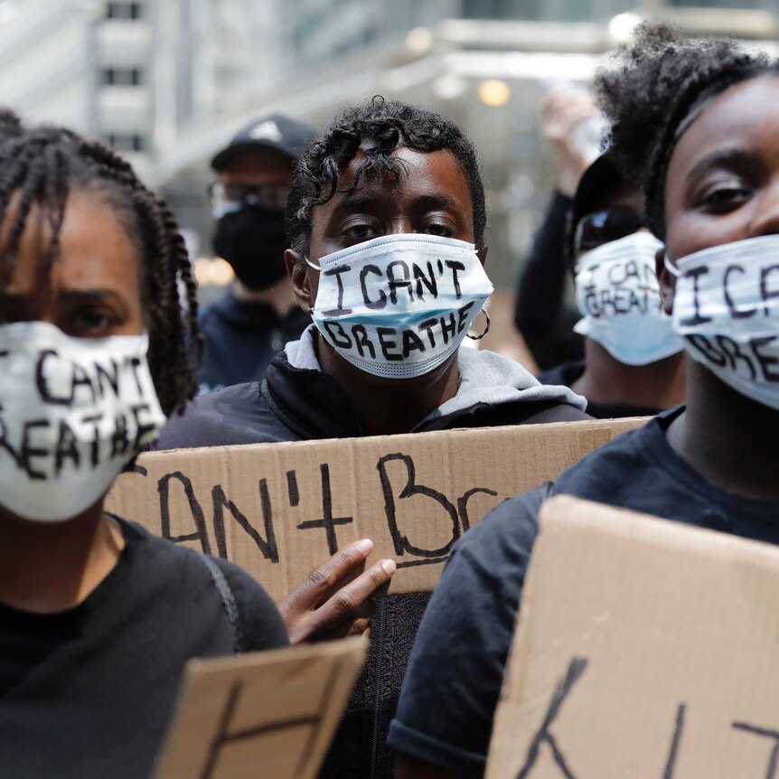 Three people stare at the camera with signs and masks reading "I can't breathe".