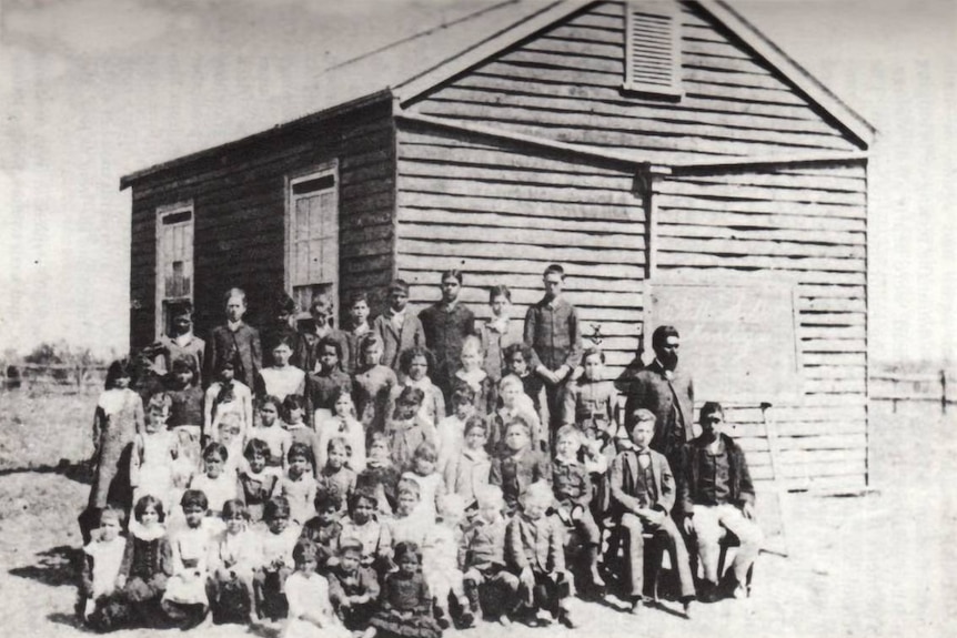 Black and white photo of about 50 children standing with a man and blackboard in front of a small wooden building.
