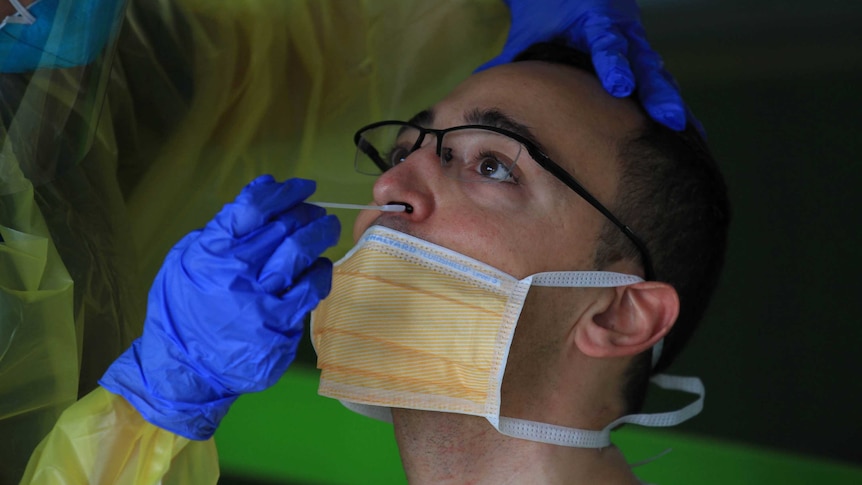 Man with glasses wearing orange facemask has plastic swab inserted in nostril by woman wearing blue gloves and yellow gown.
