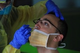 Man with glasses wearing orange facemask has plastic swab inserted in nostril by woman wearing blue gloves and yellow gown.