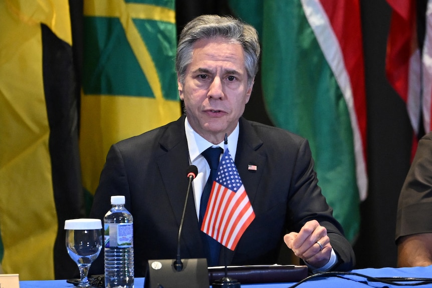 Secretary Blinken speaks into a microphone with flags behind him and the US flag in front of him