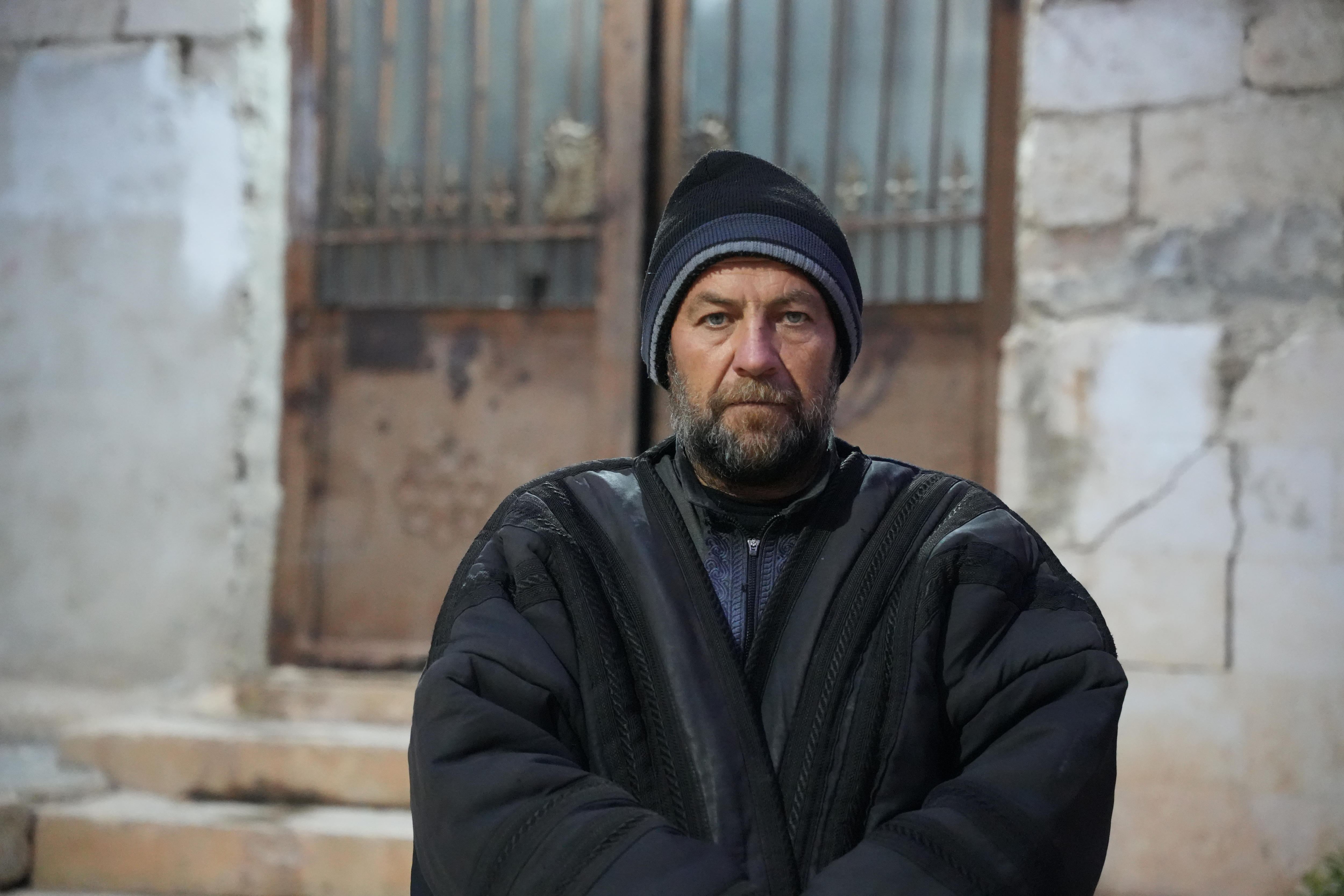 Um homem de gorro e casaco grosso com expressão solene, parado em frente a um prédio em ruínas.