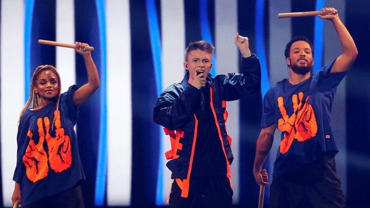 Eliot, flanked by two dancers, holds a fist up as he sings in orange and navy jacket.