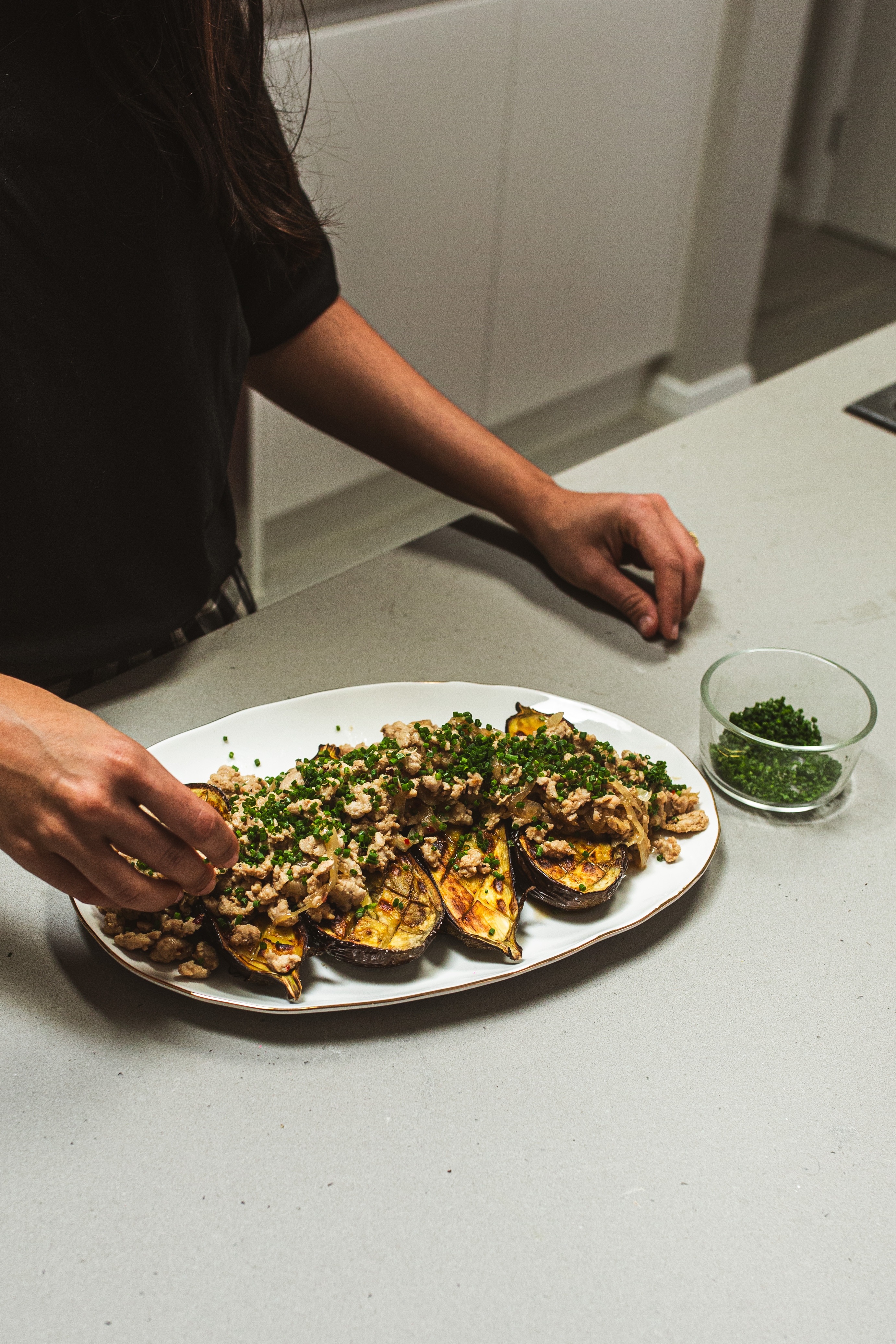 A plate of cooked eggplant halves with chicken mince on top sits on a bench. A person sprinkles chopped green chives on top.