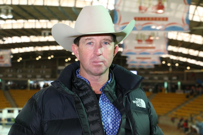 A man in a black coat and white hat stands in front of a camera.