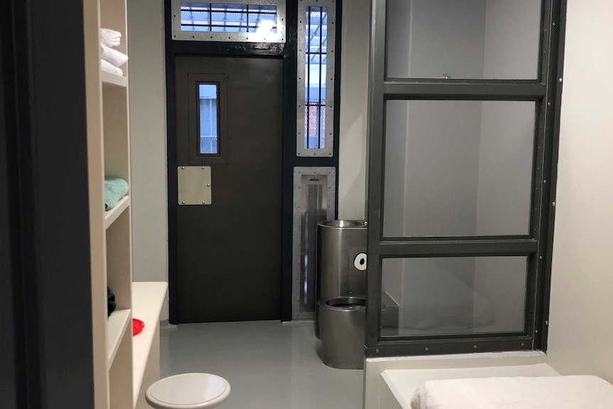 The interior of a prison cell shows a bed with a clear glass divider panels, shelves, a desk, a stool and steel sink and toilet.