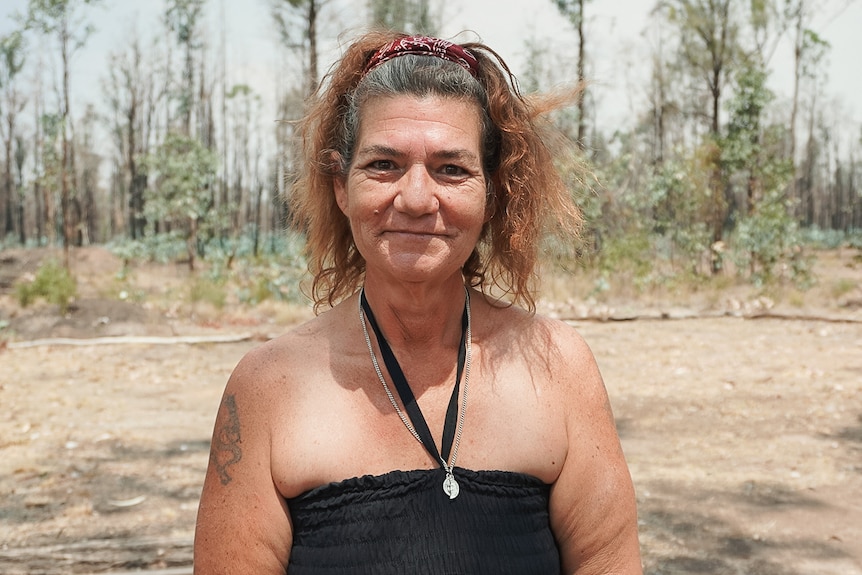 A woman stares strongly into the camera in front of burnt out trees