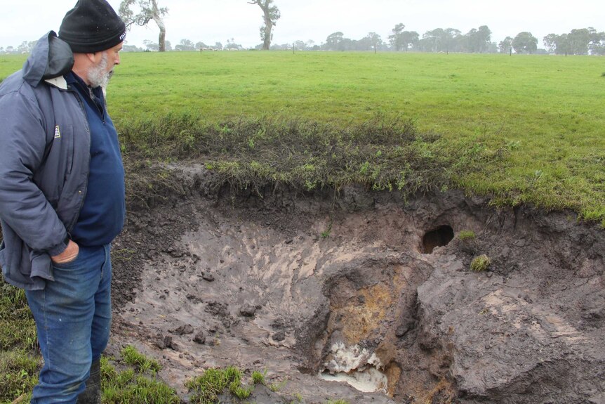 A man stands in a grassy field looking at a big hole which is flowing with water