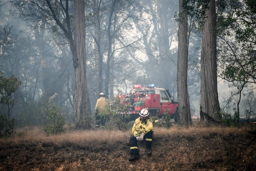 A firefighter sits exhausted on a grassy slope with a truck in the background