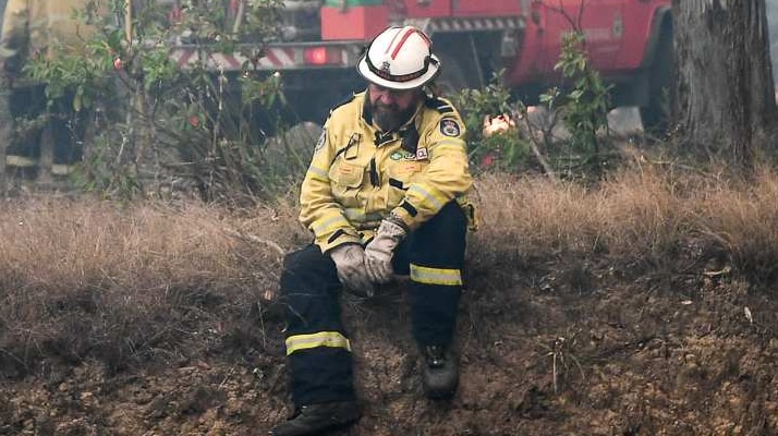 A firefighter sits exhausted on a grassy slope with a truck in the background