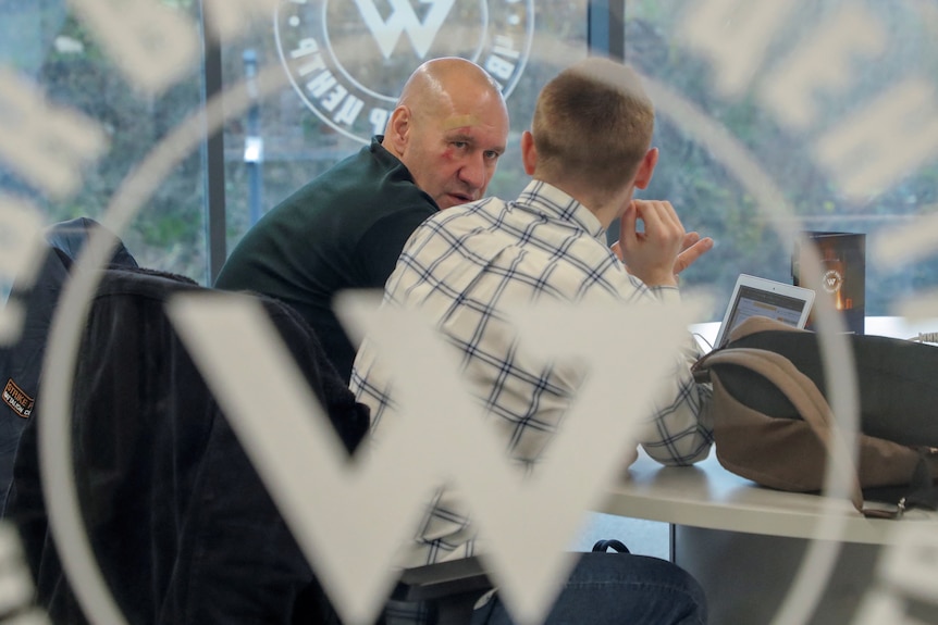 Two men - one sporting a black eye - sit at an conference desk, seen through a large W embossed on glass