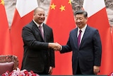A Tongan man and a Chinese man, both dressed in suits, smile and shake hands as they stand in front of their countries' flags.