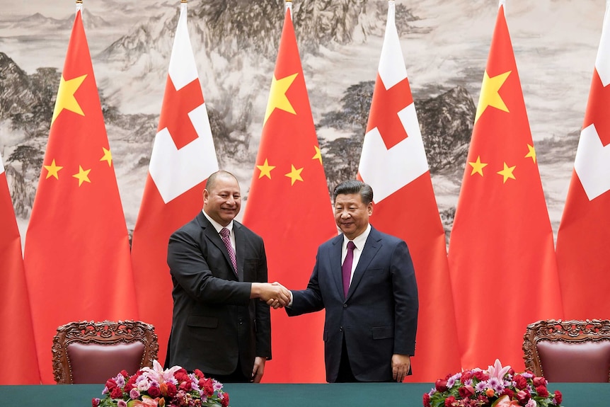 A Tongan man and a Chinese man, both dressed in suits, smile and shake hands as they stand in front of their countries' flags.