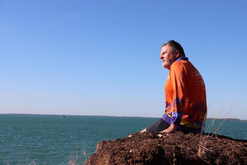 A man in an orange shirt sits on a rock and looks out to sea. 