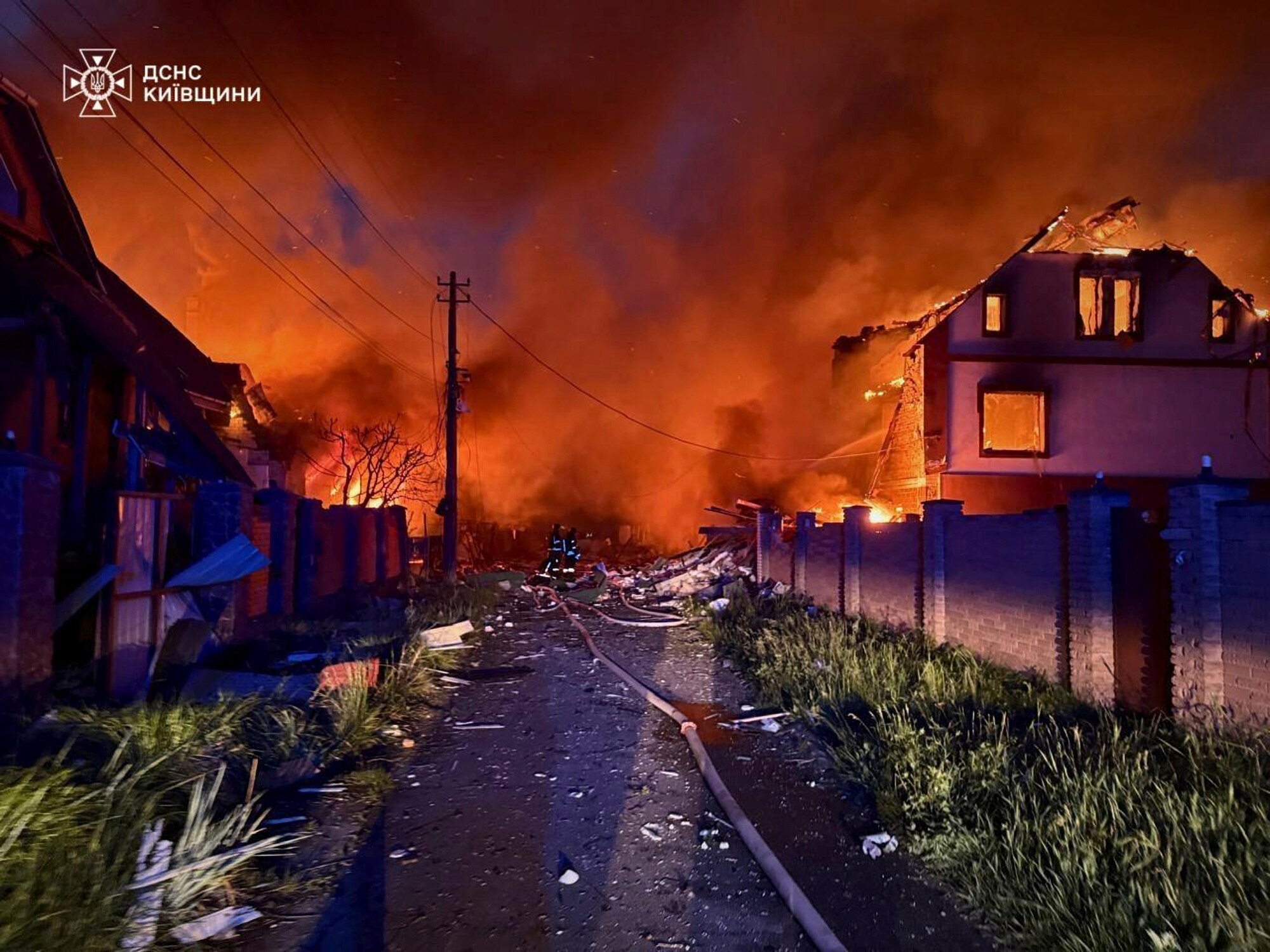 A fiery scene shows houses ablaze after drone strikes near Kyiv, with silhouetted emergency crews in the distance.