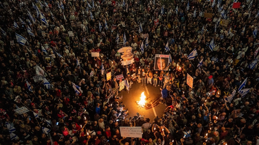 Hundreds of protesters carrying signs and Israeli flags gather around a fire