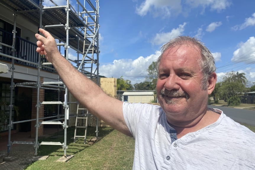 A man with grey hair and a mustache points to his home, which has scaffolding around it.