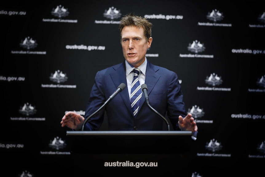 Porter is standing at a lectern in front of  Australian Government signage, both arms raised slightly.
