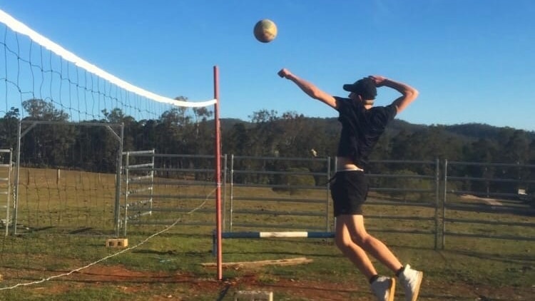 A young man in all black tee, shorts, white shoes, jumps up in the air, hits ball across net on grass, dirt, blue sky, trees.