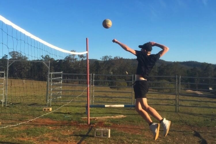 A young man in all black tee, shorts, white shoes, jumps up in the air, hits ball across net on grass, dirt, blue sky, trees.