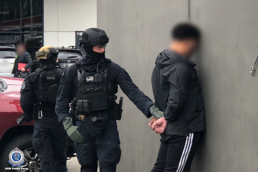 A police officer wearing helmet, balaclava, vest and gloves holds a man's hands behind his back as he faces a wall.