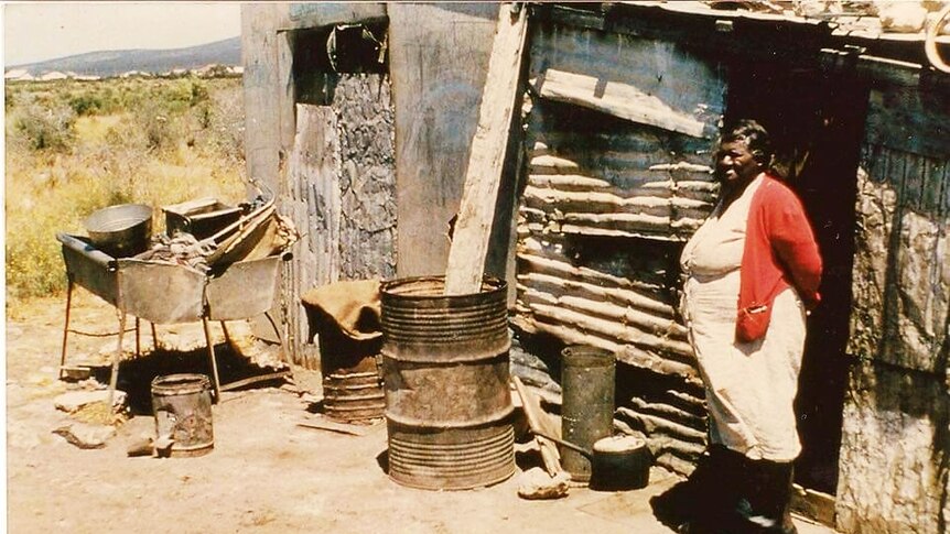 Indigenous woman standing on right in front of tin shack building