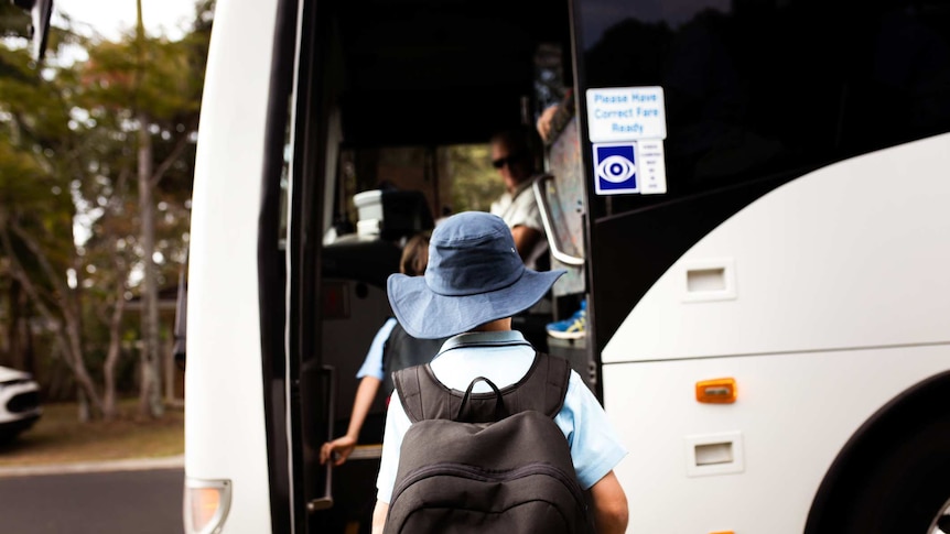 A primary school-aged student queues to get on the bus. Gum trees are in the background.