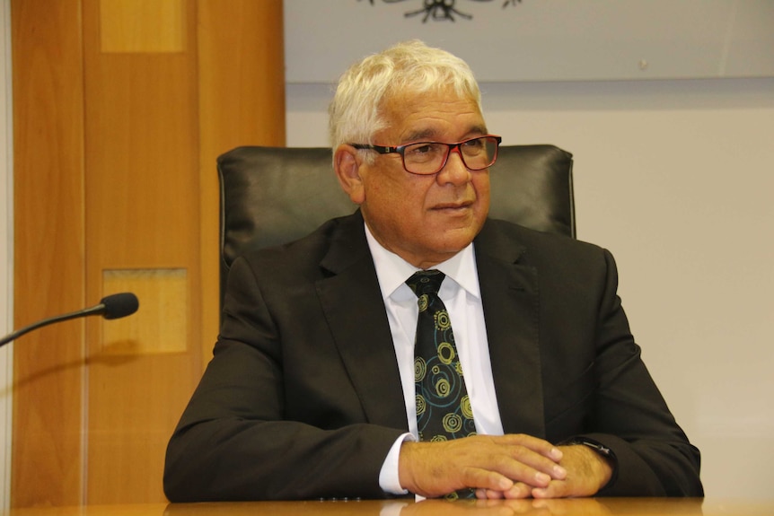 A man with white hair, in a dark suit, sits with his hands resting on the table.