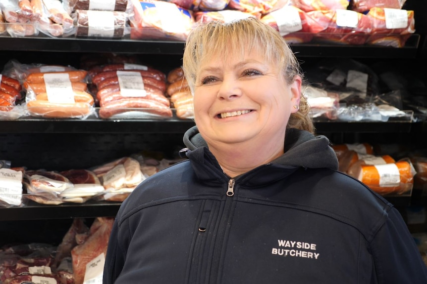 a woman in a black jumper stands in front of a fridge full of meat, she has blonde hair. 