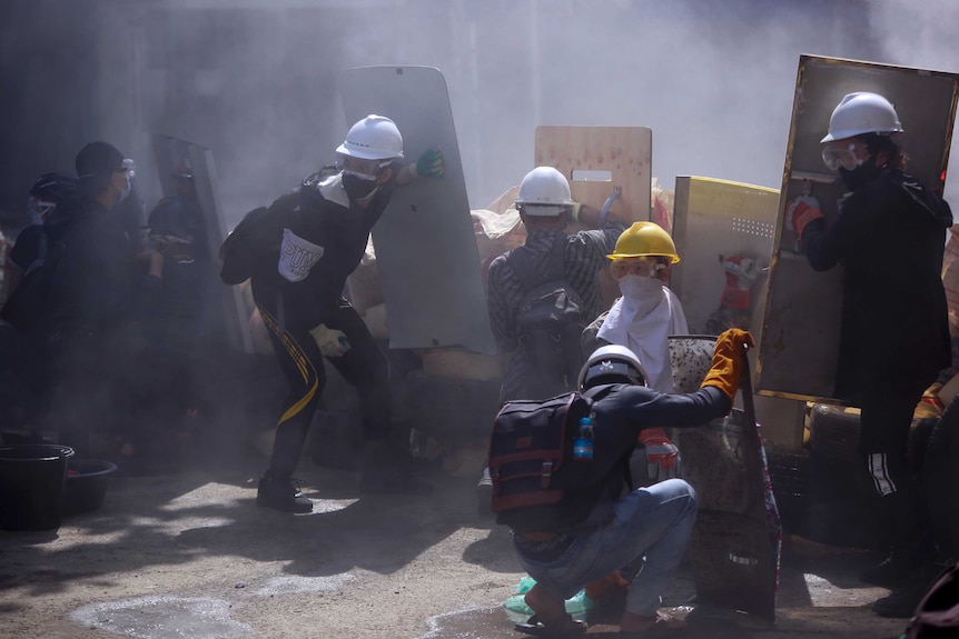 Protesters use makeshift shields and random material to make a barrier behind which they crouch as smoke billows about.