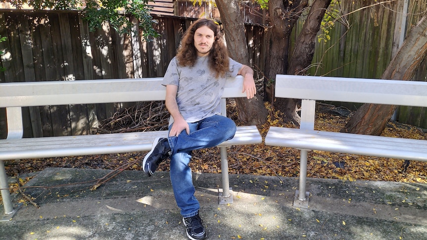 A young man sits on a silver bench in a park