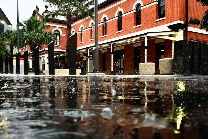 A puddle with raindrops falling into it reflects a nearby building.
