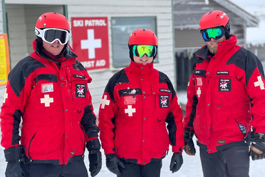 Three men wearing red snow jackets and snow goggles stand smiling in the snow.