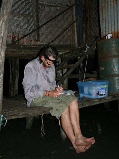 A man wearing shorts and a long-sleeved shirt with damp patches writes while sitting on a dock his legs dangling above water