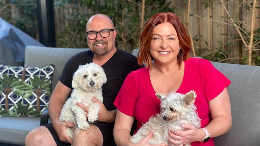 A man and woman with beaming smiles sit on their patio with little white dogs on their laps 