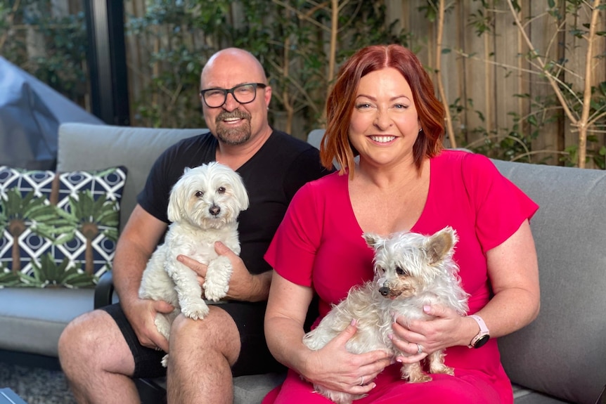 A man and woman with beaming smiles sit on their patio with little white dogs on their laps 