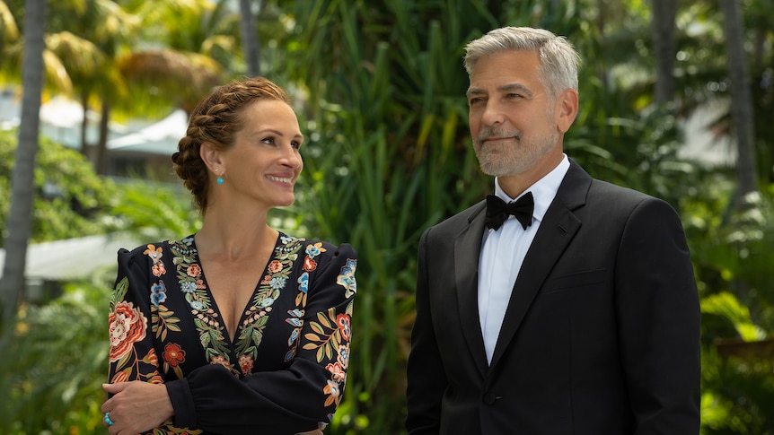 Middle-aged white woman with red braided hair wears floral black dress beside grey-haired man in black tuxedo.