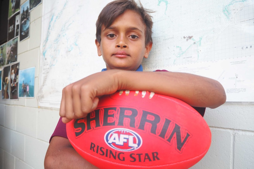 A photo of a young boy with an AFL ball staring at the camera