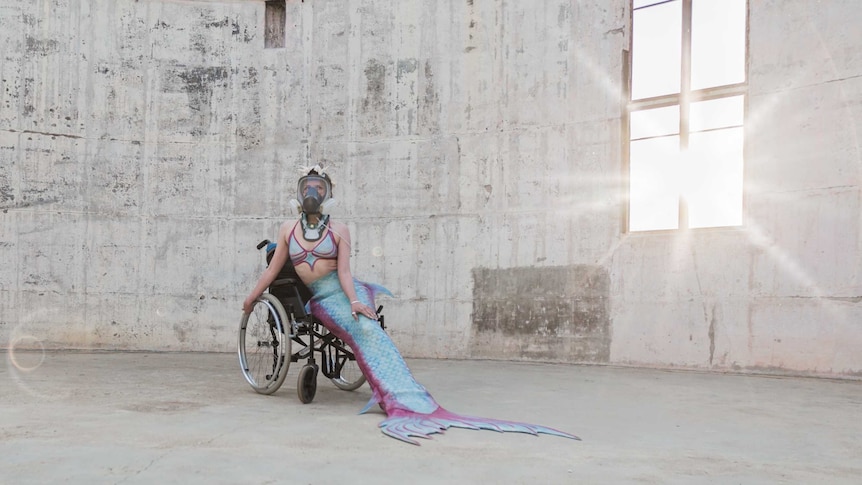 Hanna Cormick dressed as a mermaid with a long tail and wearing a respirator mask sits in a wheelchair in a large concrete room.